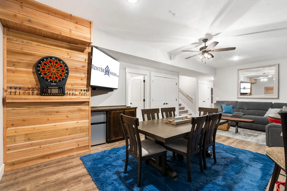A cozy, casual living area featuring a dartboard, dining table, blue rug, sofa, and TV screen. The setting is perfect for laid-back family dinners, watching movies, or having a game night.