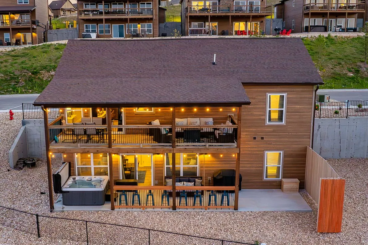 The image showcases the expansive deck of a single-family house, featuring a private hot tub, comfortable outdoor seating, and glowing porch lights. The deck offers stunning sunset views over the lake and is perfectly set up for relaxing, dining, and spending time with loved ones in a serene mountain setting.