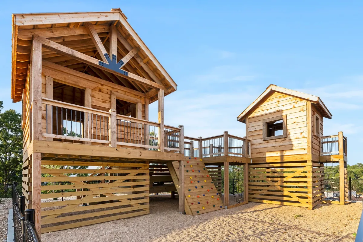 An exterior shot focusing on a charming wooden playhouse equipped with a climbing wall, slide, and stairs, set outdoors. Children are actively playing, climbing, and exploring, radiating joy and adventure. The scene embodies imaginative, active play for kids and the family-friendly aspect of the resort.