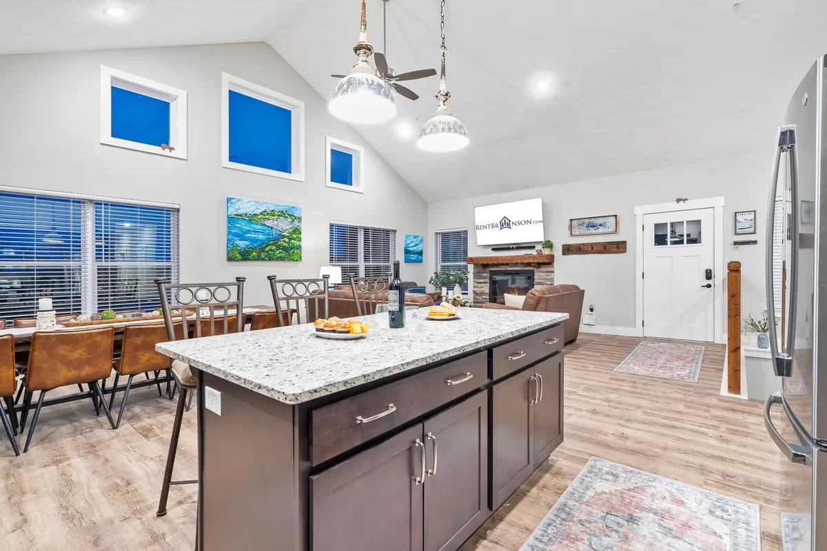 An open-concept space that beautifully connects the kitchen, dining area, and living room. The kitchen island and dining table are visible, with stylish bar stools and modern artwork, inviting guests to gather, cook, dine, and relax together in a sociable setting.