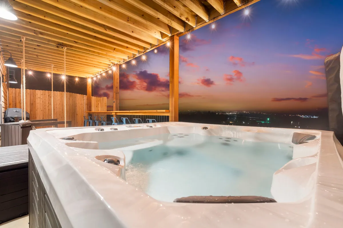 A luxurious outdoor hot tub sits under a wooden ceiling adorned with string lights, facing a vivid sunset sky. The ambiance is tranquil and romantic, inviting guests to relax and socialize while surrounded by natural beauty.