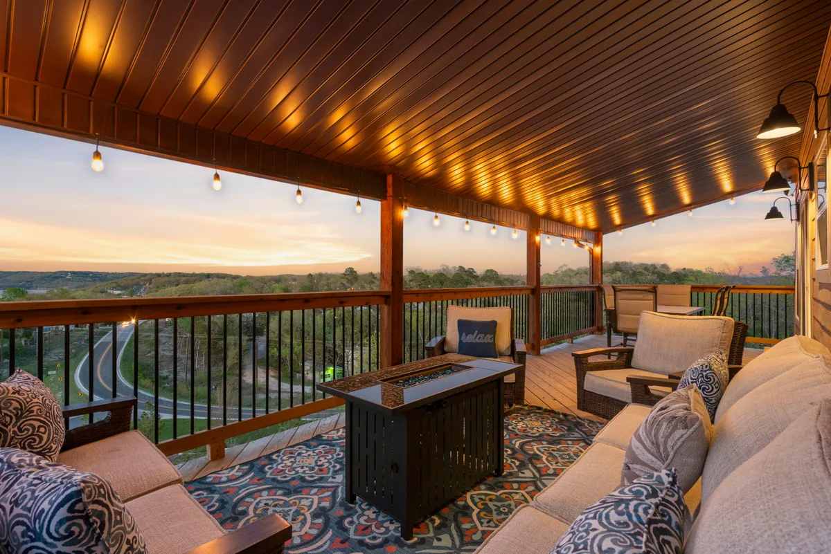 An outdoor deck area featuring comfortable seating, a decorative rug, string lights, a fire pit, and expansive views of the surrounding landscape. The warm glow and open-air ambiance make it perfect for sunset toasts, outdoor dining, and gathering by the fire with a backdrop of the Kimberling City Bridge.