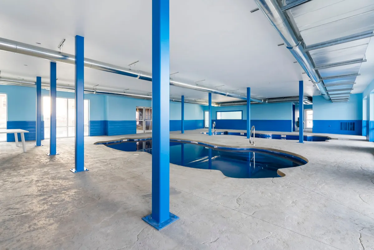 A tranquil interior view of a spacious indoor swimming pool area, featuring blue columns, large windows that let in natural light, and poolside seating. The atmosphere is inviting and serene, perfect for swimming or relaxing regardless of weather, and conducive to year-round recreation.