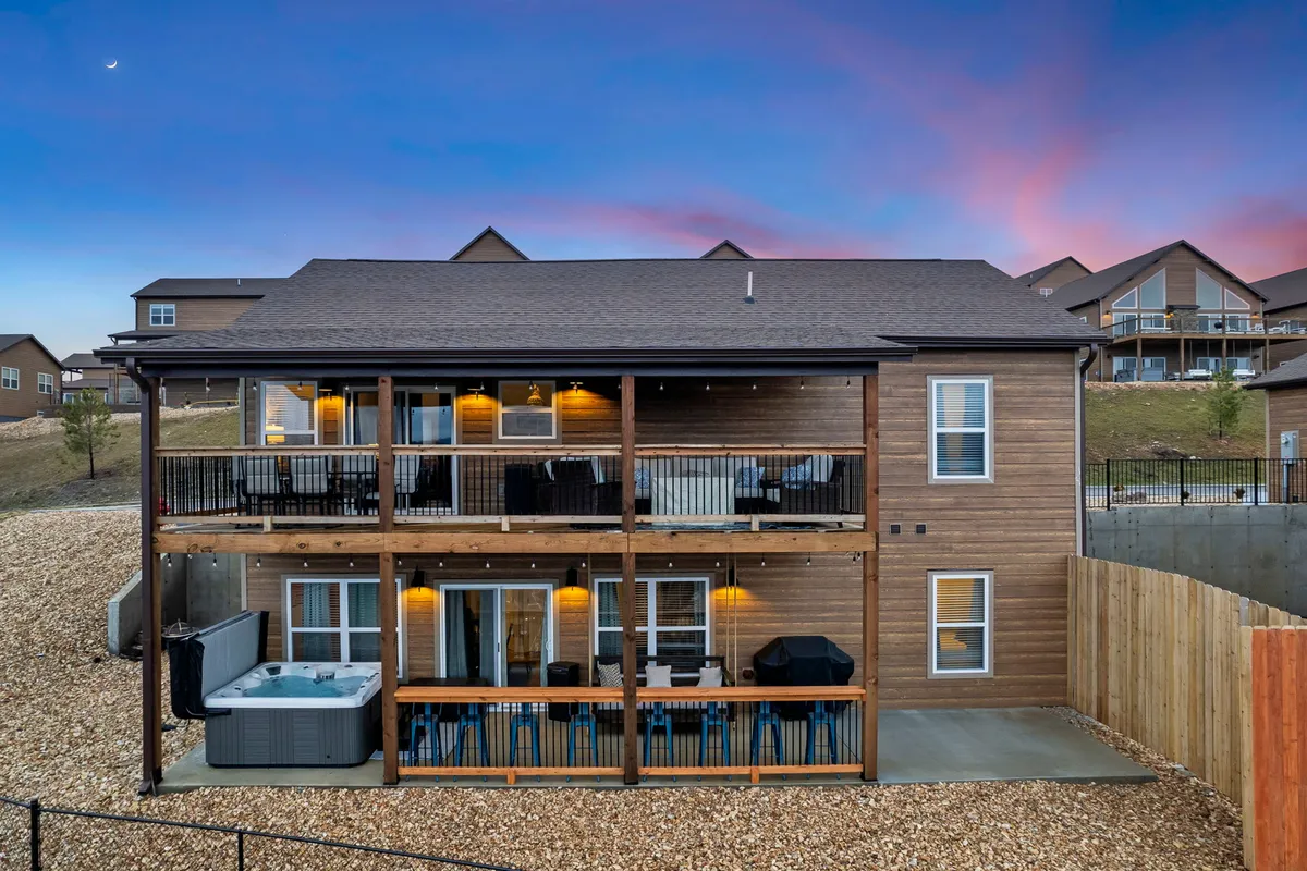 A spacious two-story wooden deck featuring a hot tub, cozy outdoor seating, and warm string lights. The deck overlooks a stunning sunset landscape, blending comfort with the natural beauty of the surroundings.